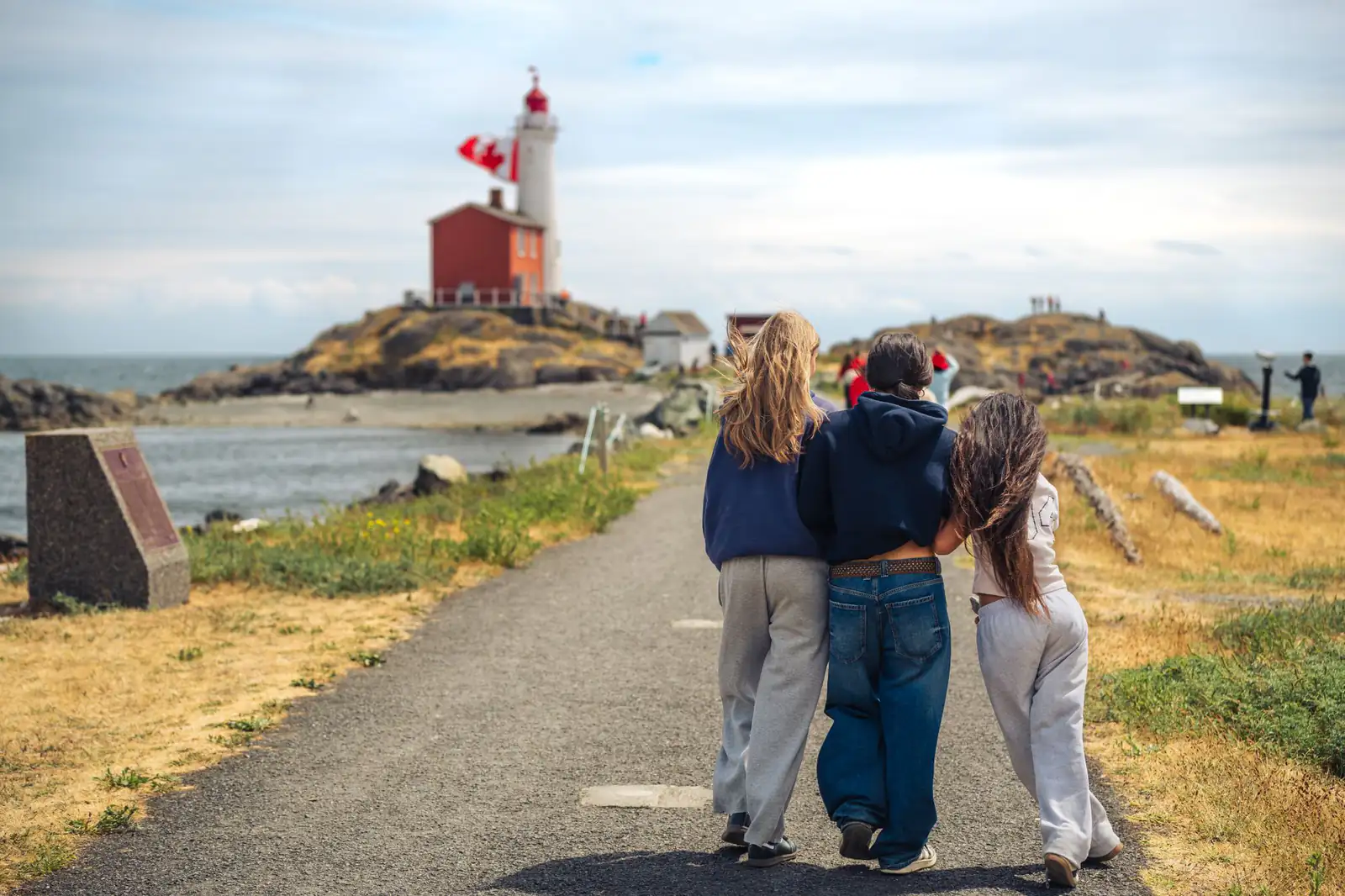 Three friends walking toward a lighthouse along a waterfront path in Victoria